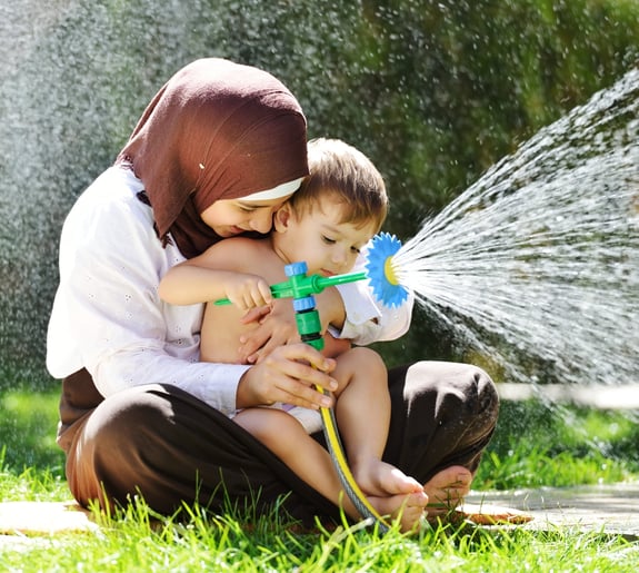 Beautiful Middle Eastern Arabic girl having baby playing with water sprinkler in garden