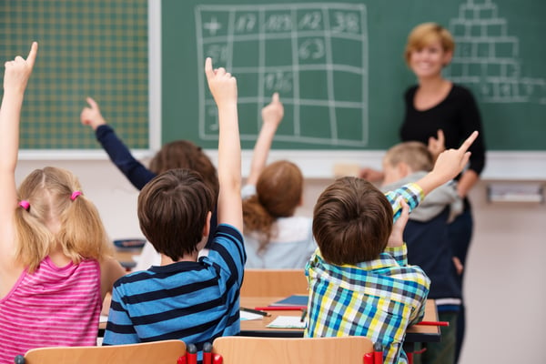 Intelligent group of young school children all raising their hands in the air to answer a question posed by the female teacher, view from behind