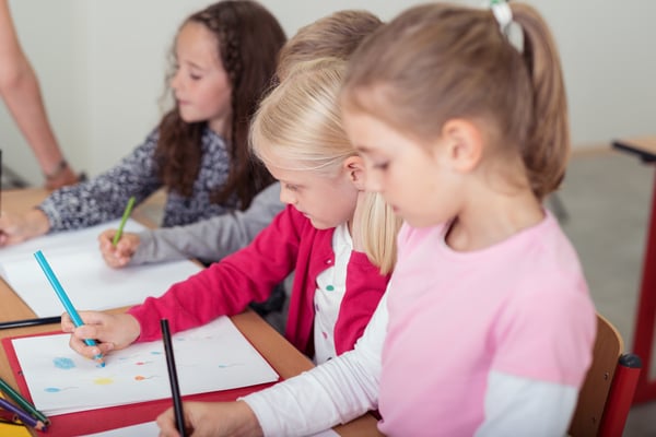 Row of small children in art class at school sitting at a long desk drawing with colored pencil crayons, focus to a young girl in the foreground